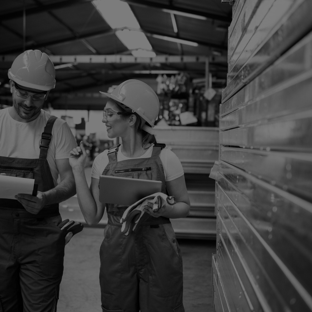 Two people wearing hard hats and overalls are discussing work in an industrial setting, possibly a warehouse, with metal materials in the background.