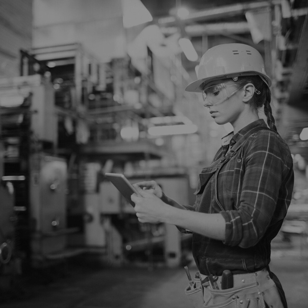 A person wearing a hard hat and safety glasses is holding a tablet, with a blurred industrial background suggesting a factory or workshop setting.