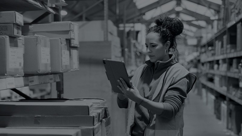 A person in a warehouse is focused on a digital tablet, possibly managing inventory. They are wearing casual work clothes and the image is monochrome.