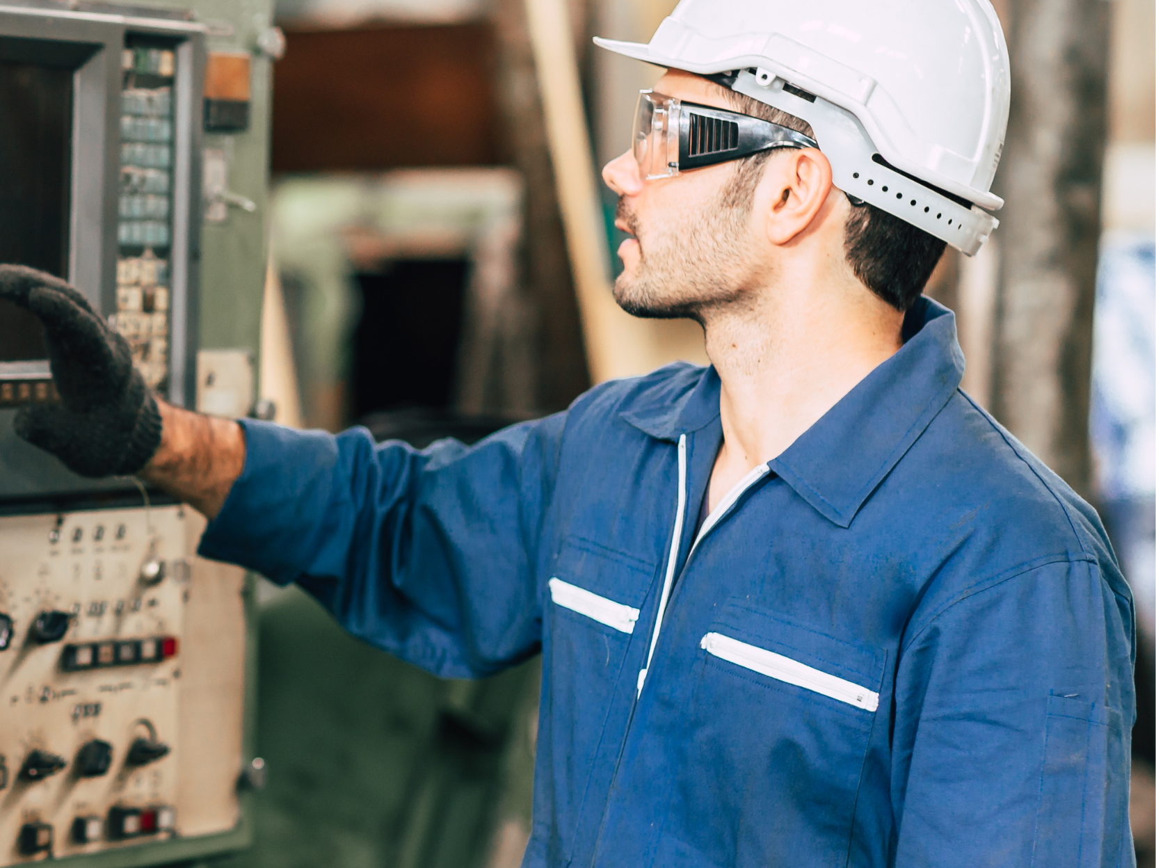 A person in a blue uniform and white hard hat is operating machinery, wearing safety glasses and a glove, in an industrial setting.