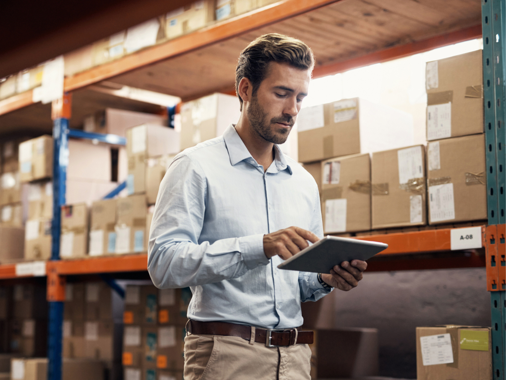 A person in a warehouse is focused on a tablet. They are wearing a blue shirt and beige pants, standing beside shelves stacked with boxes.