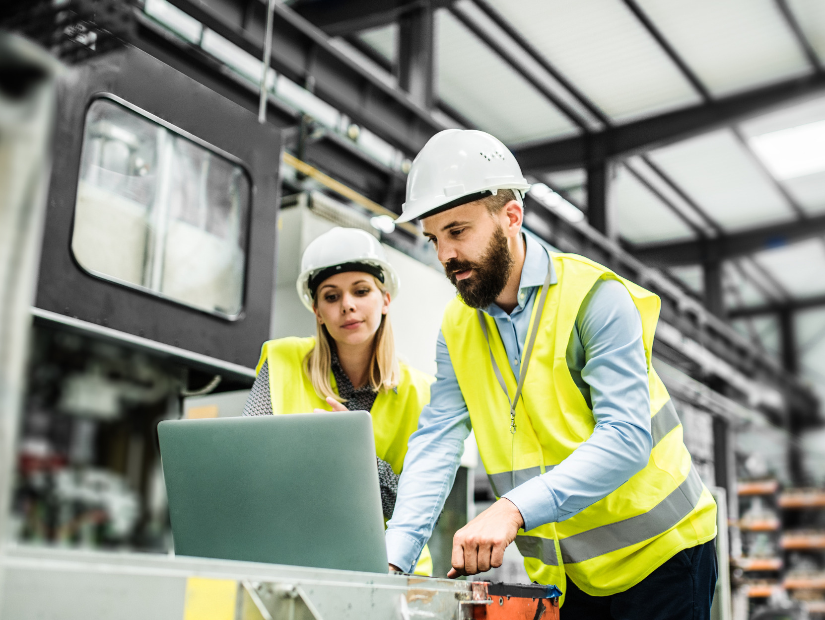 Two people wearing hard hats and high-visibility vests are focused on a laptop in an industrial setting, possibly discussing work-related matters.
